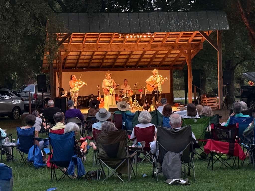 The four of them on an open timber stage at dusk, string lights overhead and the audience out on the grass in camp chairs