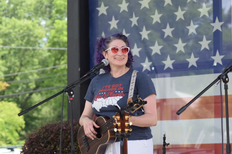 Kristi Casey at a microphone with her guitar in front of a large American flag
