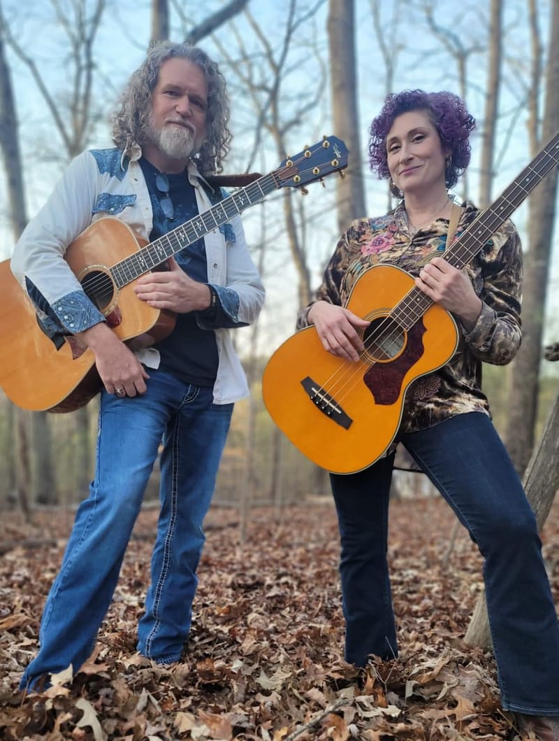 Kristi and Steven standing in the woods with their guitars, autumn leaves underfoot
